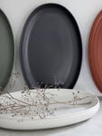 A collection of oval ceramic plates in muted tones, including green, black, and terracotta, neatly arranged upright on a shelf. In front, two white plates lie flat with delicate, dried branches placed on them.