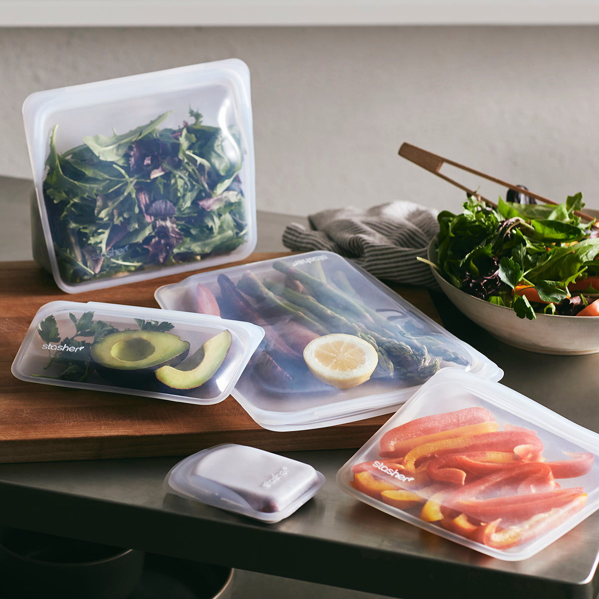 Reusable silicone bags of various sizes are filled with fresh produce, including leafy greens, sliced peppers, an avocado, and a lemon half. The bags are displayed on a wooden cutting board on a kitchen counter next to a bowl of salad.