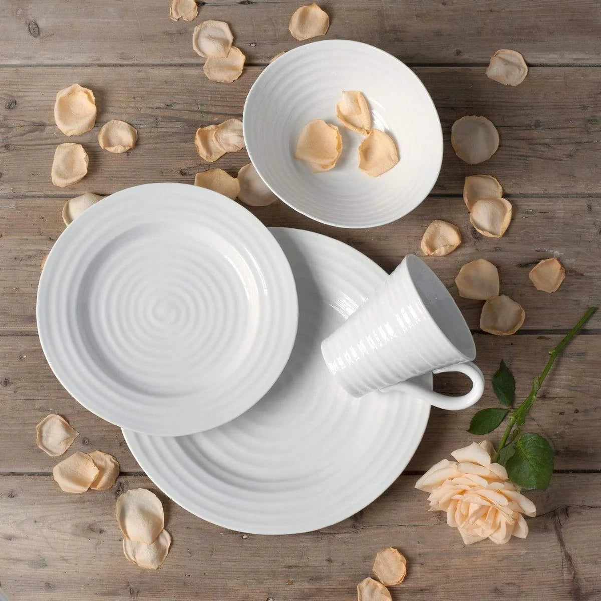 A rustic table setting featuring a white ceramic dinner set, including a plate, bowl, saucer, and cup arranged on a wooden table. Peach rose petals are scattered around, with a fresh peach rose and stem on the right.