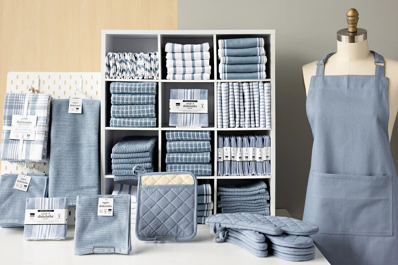 A display of kitchen textiles in hues of blue and white, including towels, dishcloths, potholders, and an apron. They are neatly arranged in a white cubby shelf and laid out on the table for viewing.