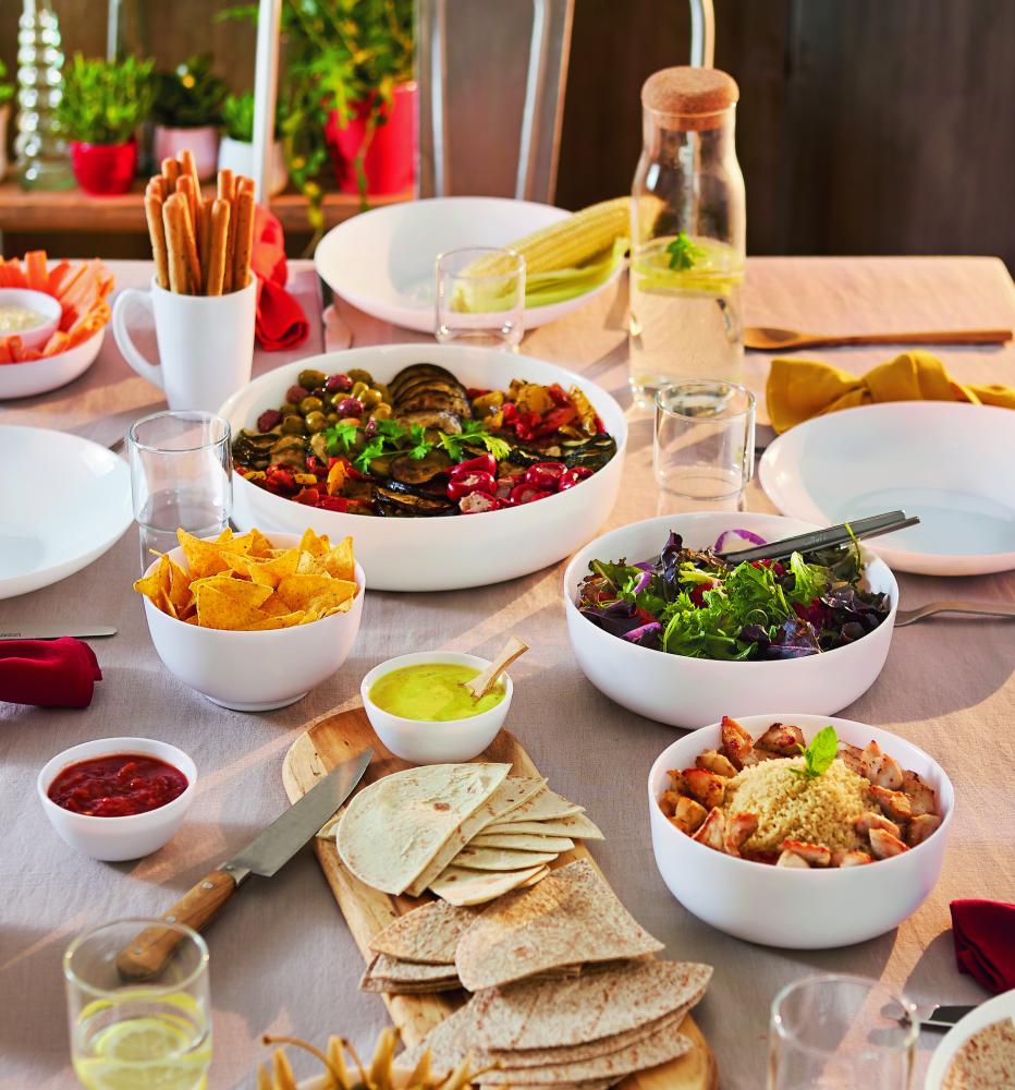 A dining table is set with various dishes including a large bowl of grilled vegetables, a bowl of nachos with salsa, a bowl of mixed greens salad, wraps, diced chicken, guacamole, breadsticks, and drinks.