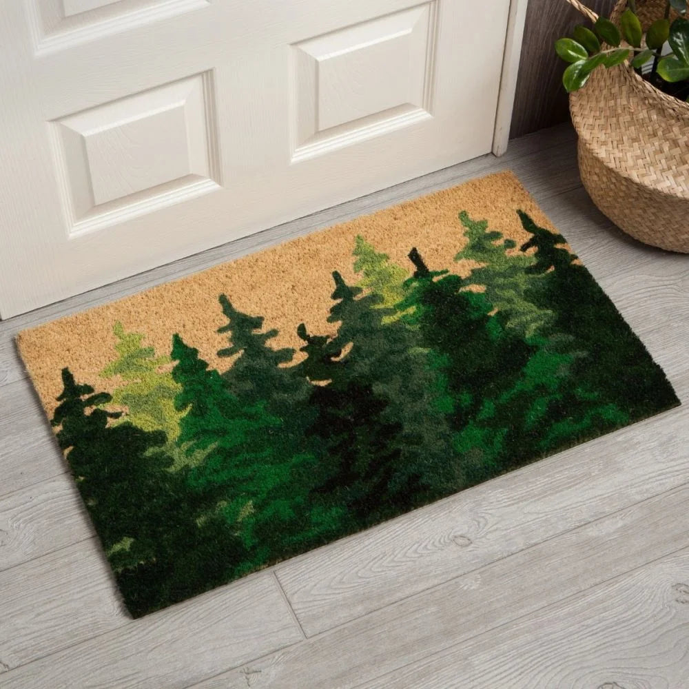 A rectangular doormat with a design featuring a dense green forest against a beige background, placed in front of a white door on a light gray wooden floor. A woven basket is partially visible on the right.