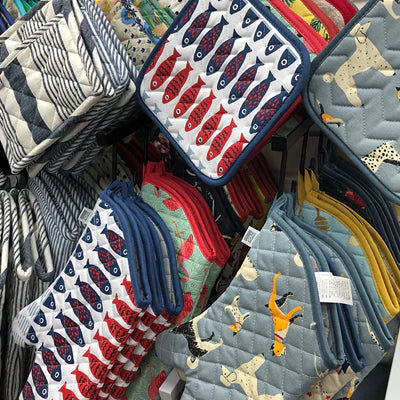 Several patterned oven mitts and pot holders are displayed on hooks in a store. Designs include striped, fish, and animal themes in various colors like blue, red, and gray.