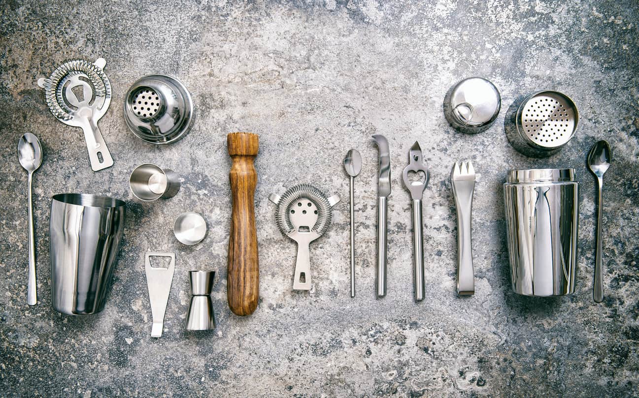 Various cocktail making tools neatly arranged on a textured surface, including shakers, strainers, spoons, a wooden muddler, jiggers, a bottle opener, and mixing utensils, all in metallic tones.