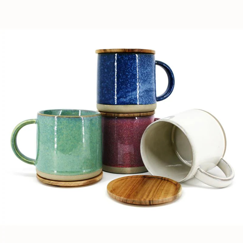 Four ceramic mugs are displayed, each with a wooden lid. The mugs are in blue, green, red, and white, with the blue and red mugs stacked and the green and white mugs lying on their sides. The mugs have a textured design.