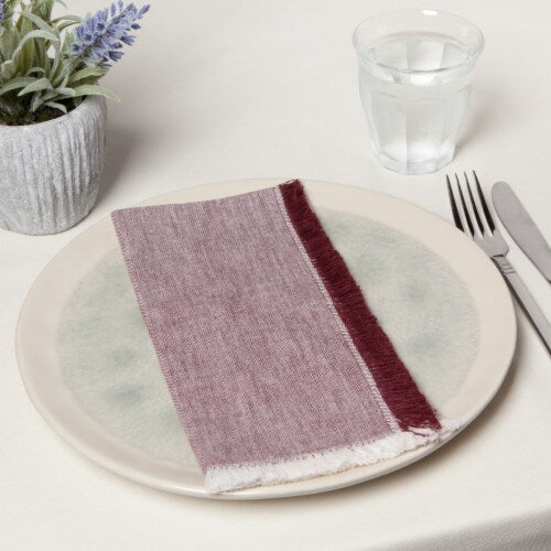 A neatly set table with a white plate, a burgundy and purple cloth napkin, and a fork and knife to the right. A glass of water and a potted plant are in the background.