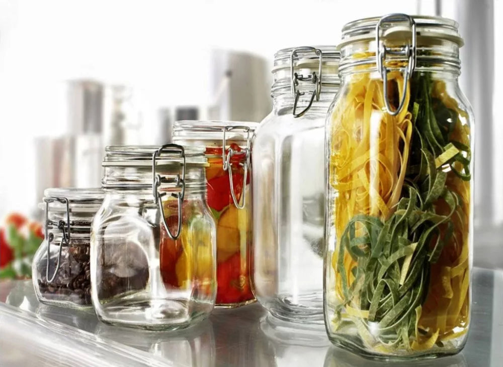 Various sizes of glass jars with metal clasps are filled with food items, including colorful dried pasta, pickled vegetables, and coffee beans, arranged on a reflective surface. The background is blurred, highlighting the jars.