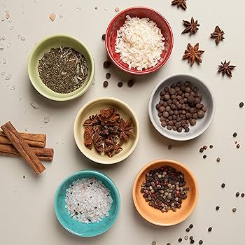 Top view of six small bowls containing various spices: dried herbs, rice, allspice, star anise, coarse salt, and peppercorns. Cinnamon sticks and scattered star anise and peppercorns are also visible on the light-colored surface.