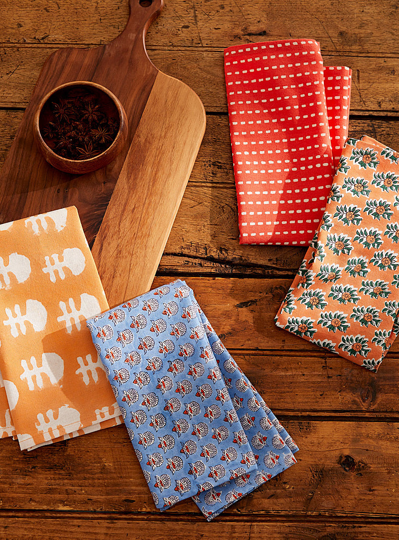 Four colorful patterned kitchen towels are neatly folded on a rustic wooden table. Next to them, a wooden cutting board with a small bowl containing star anise is seen. The towels feature various designs in orange, blue, and red tones.