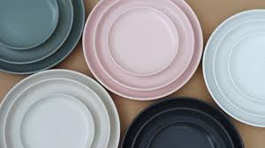 A collection of ceramic plates in shades of green, pink, white, gray, and black, arranged in a semi-circle on a neutral-colored background. Each set includes a large and a smaller plate, stacked together.