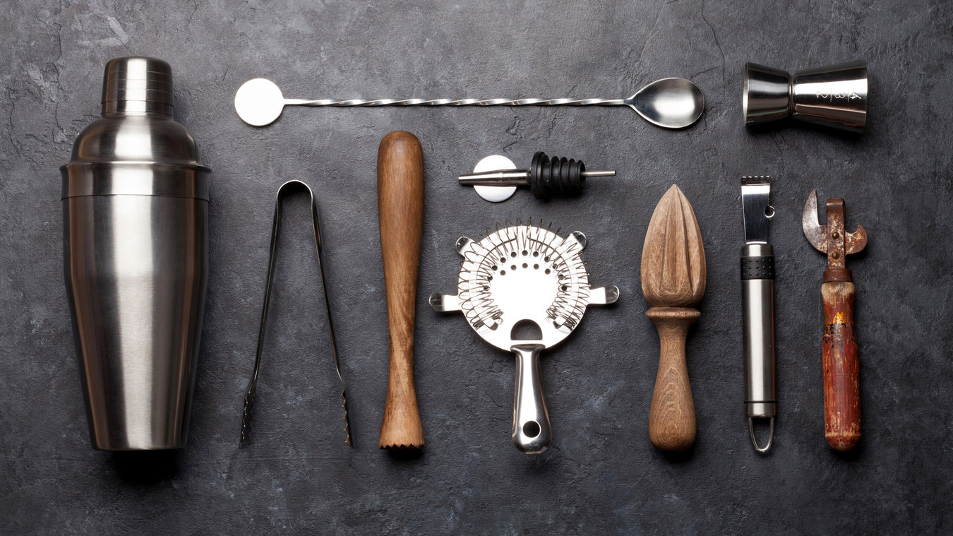 Top view of various cocktail tools on a dark textured surface, including a shaker, mixing spoon, double jigger, ice tongs, muddler, pour spout, strainer, wooden reamer, peeler, and can opener.