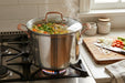 A large stainless steel stockpot with copper-toned handles sits on a gas stovetop in a kitchen, with steam rising from soup inside. A wooden cutting board with chopped onions, carrots, and celery is visible on the counter to the right.